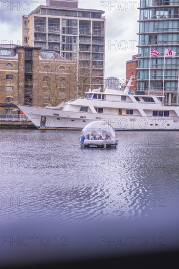 A dome boat on calm water in front of a modern marina with buildings in the background, London,