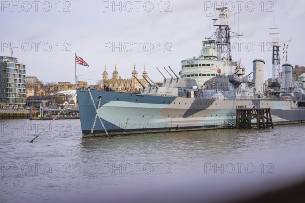 Historic warship on the riverbank in a city with British flag and buildings in the background, HMS