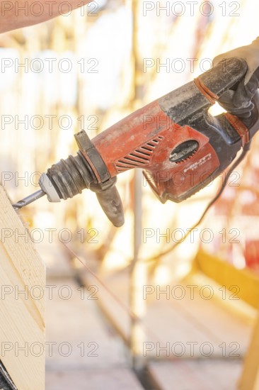 A construction worker operates a red drill on a construction site. Sunlight illuminates the scene,