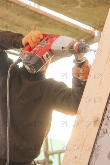 A worker uses a drill to process a slanted wooden board under sunlight, carpentry construction,