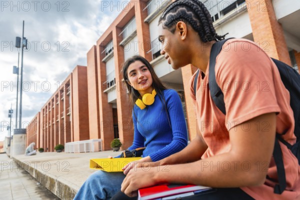 Two diverse young students engaging in a friendly conversation while sitting on a ledge on the