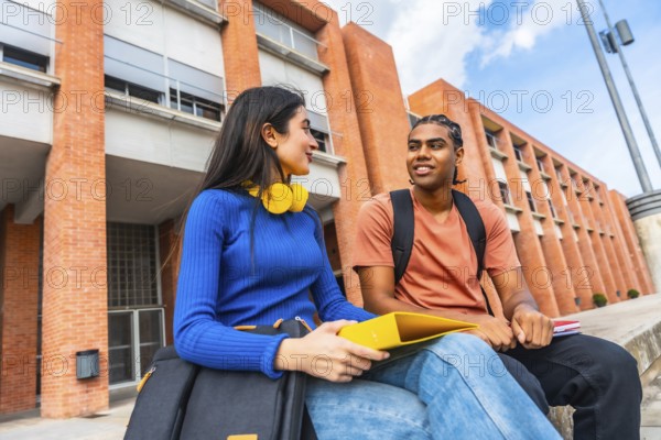 Two young diverse students are sitting on a wall outside a university building, engaging in a