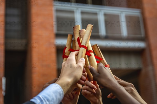 Graduates from various backgrounds raise their rolled up diplomas, tied with red ribbons,