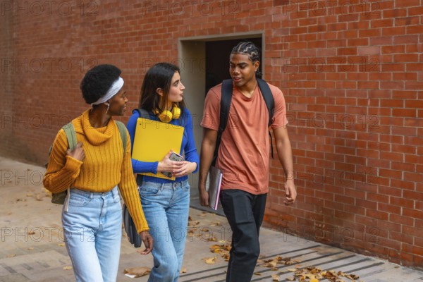 Three diverse young university students walking along a brick wall, carrying backpacks and books,