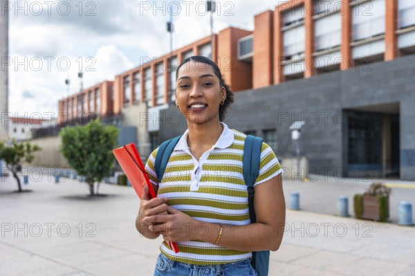 Young female student smiling while holding a folder and wearing a backpack, standing outdoors on a