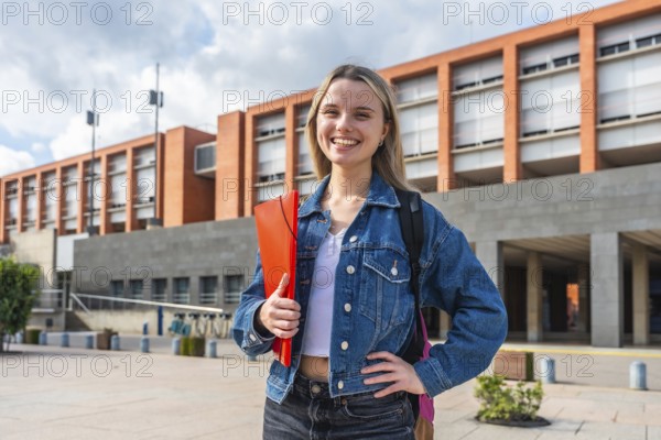 Happy young female student with backpack and folder smiling, standing in front of a modern