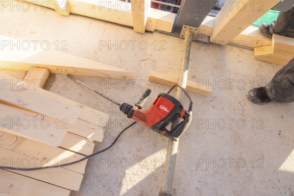 A drill is lying on a construction site floor, surrounded by wooden planks and a pair of shoes,