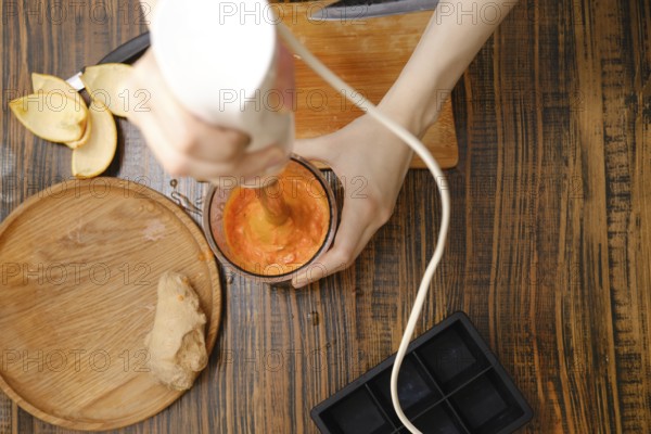 A person uses a hand blender to mix fruits and ginger in a cup. The setting looks like a kitchen
