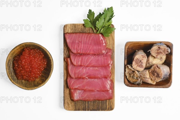 Sliced cured tuna fillets and canned tuna are on a wooden board, accompanied by a bowl of red fish