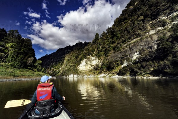 Person in a canoe paddles along steep, wooded cliffs under a dramatic sky, zero