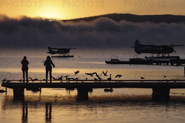 A seaplane on Lake Rotorua at sunrise, with silhouettes of people and birds on the dock, Rotorua,