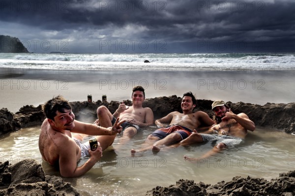 Group of men relaxing in waterhole on beach under dramatic clouds, Hot Water Beach, Coromandel