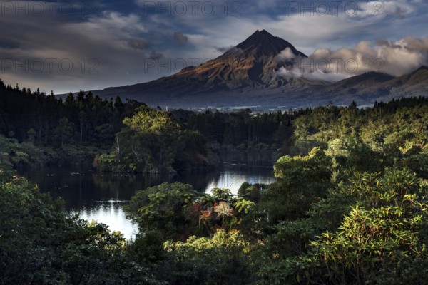 Mount Taranaki rises majestically above Lake Mangamahoe surrounded by lush greenery, New Zealand,