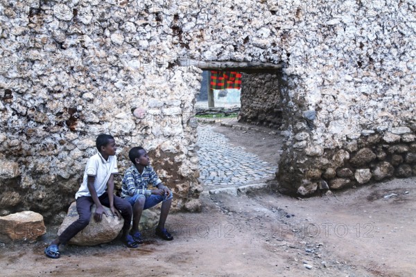 Two boys sit in front of a hyena gate in an old stone wall in the old town of Harar, Harar, Harar,