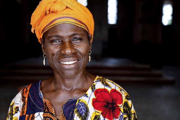 Woman in colorful dress with yellow headscarf smiles at the camera. Dark background, Bouaké,