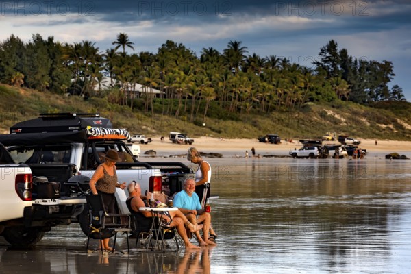 Visitors relax next to cars on Cable Beach car beach, Broome against a palm-covered background,