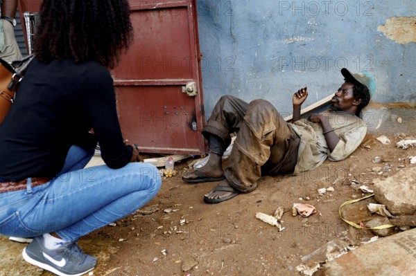 Mentally ill homeless person sits in front of a door. A woman approaches him on a dusty road,