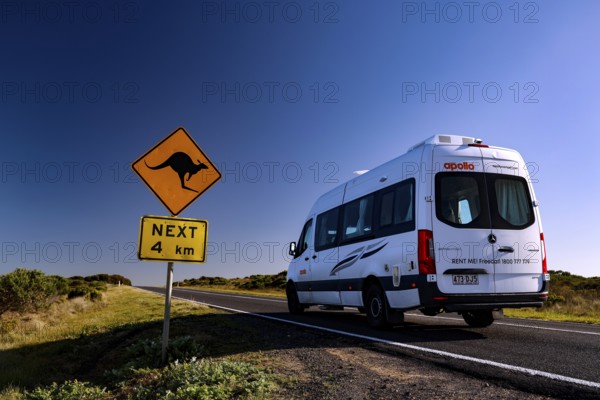 A motorhome drives along the Great Ocean Road past a kangaroo warning sign under clear blue skies,