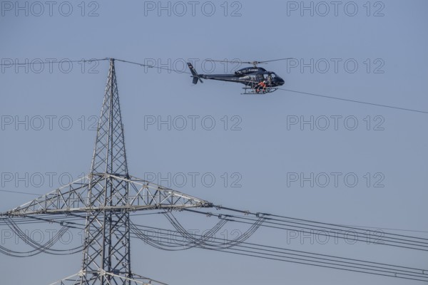 A helicopter carries out technical work on power lines next to a mast under a blue sky, an employee