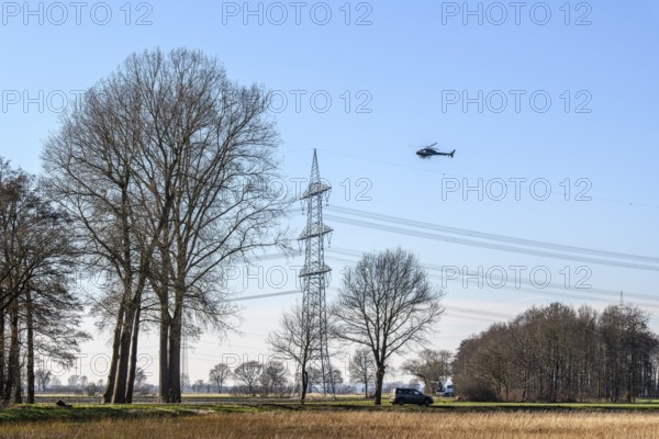 A helicopter flies over a landscape with power poles and trees under a blue sky, Lemförde, Germany,