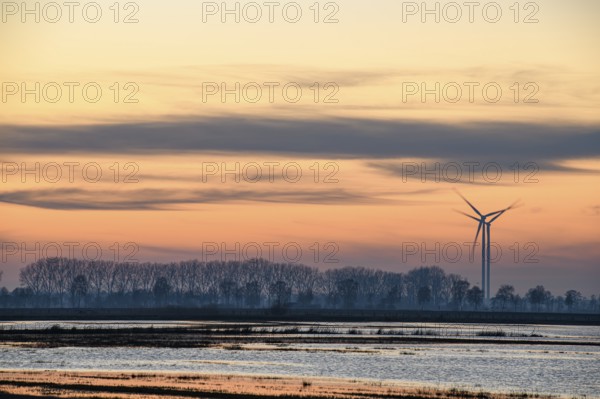 A wind turbine at sunset next to a calm body of water and trees, wind turbines in front of an