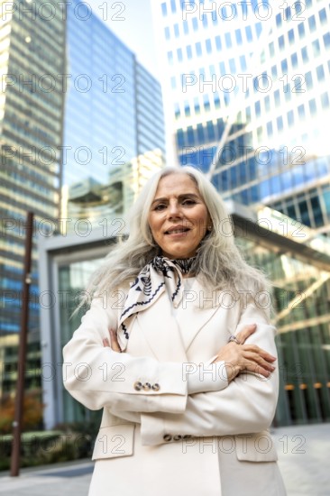 Senior businesswoman with gray hair in a cream suit stands confidently with arms crossed before
