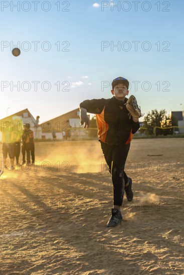 Young boy in baseball uniform pitching a baseball, creating a cloud of dust on the field during an
