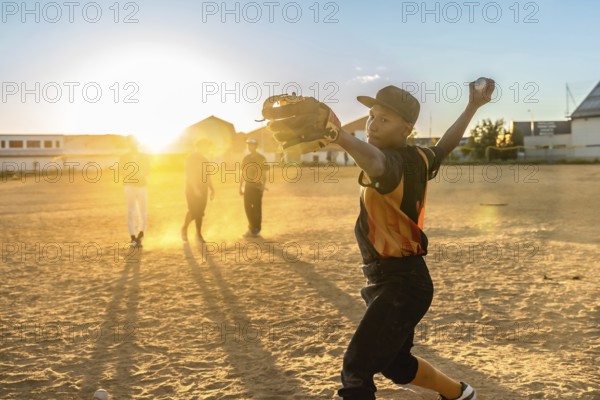 Boy pitcher throws a baseball on a dusty summer field at golden hour, practicing with teammates,