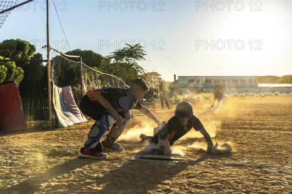 Young baseball player sliding into base on dusty infield as catcher reaches to tag him, dynamic