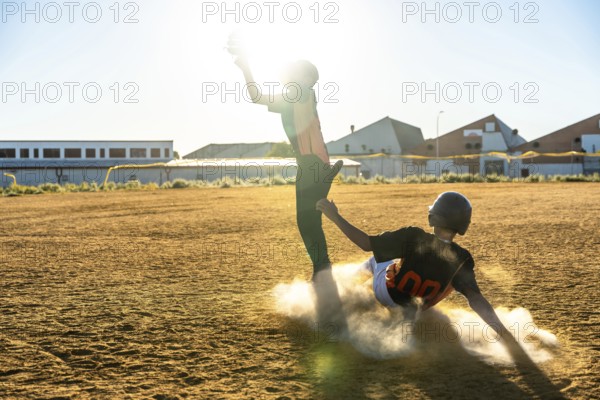 Two young baseball players are competing on a dusty field, one sliding vigorously into a base and