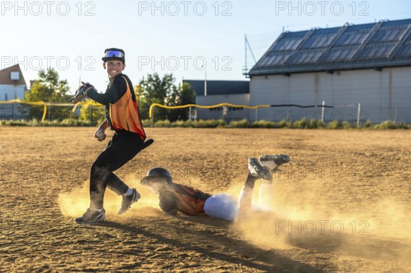 Young baseball players competing on a dusty field, one sliding head first into a base while an