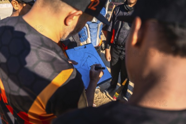 Baseball coach in uniform drawing game strategy on clipboard while explaining plays to youth team