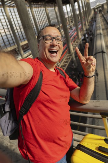 Middle aged man wearing reading glasses smiles with teeth while taking a selfie and making a peace