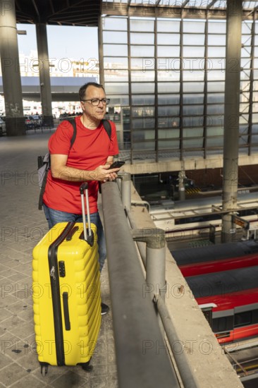 Middle aged man standing at a city train station with a yellow suitcase and looking at the railway