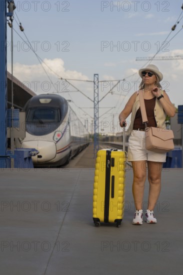 Adult woman walking on a modern train platform with a yellow suitcase, waiting for departure. Solo