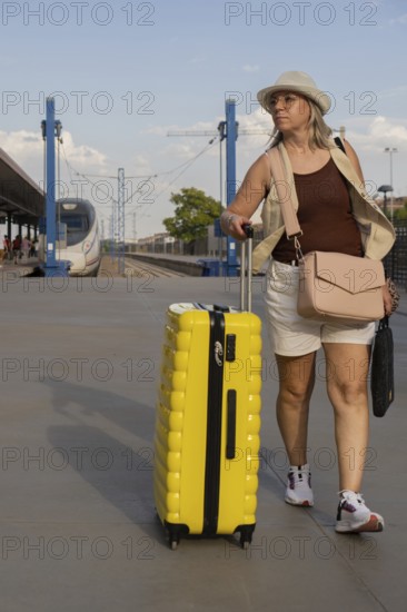 Adult woman traveling alone at a city train station, pulling a yellow suitcase while walking along