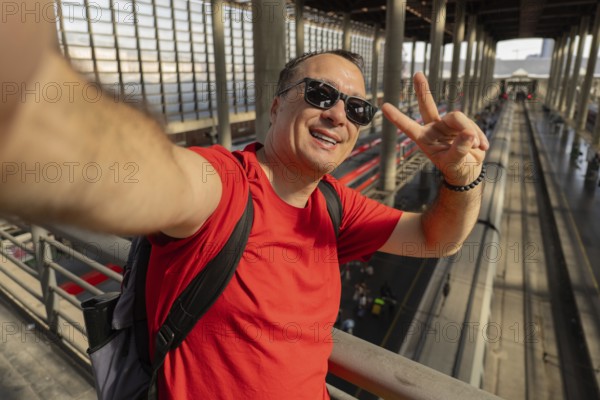 Middle aged man wearing sunglasses takes a cheerful selfie at a busy train station. Modern travel,