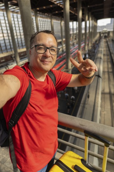 Middle aged man wearing reading glasses smiles calmly while taking a selfie and making a peace sign