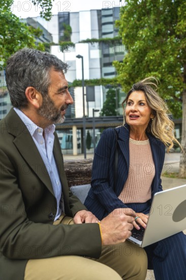 Two professional business colleagues, a man and a woman, are sitting on a bench in a city