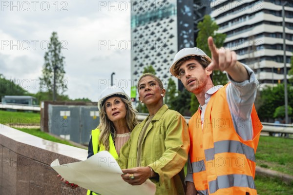 Diverse construction team in hard hats and safety vests reviews blueprints and points toward an