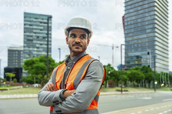 Young man wearing a hard hat and safety vest standing confidently with arms crossed in a cityscape,