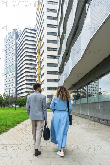 Two business professionals walking together along a paved pathway in an urban area, passing tall