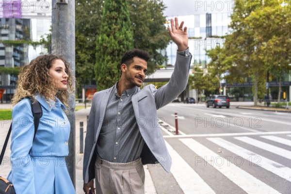 Young businessman hails a taxi on a busy downtown sidewalk while his female colleague stands beside