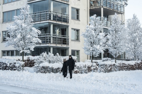 A couple walks on a snow-covered street in front of apartment buildings and snow-laden trees in