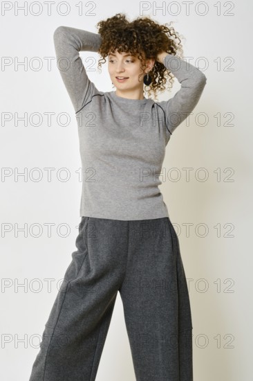 A woman stands with her hands behind her head, showcasing a cozy grey long-sleeve top paired with