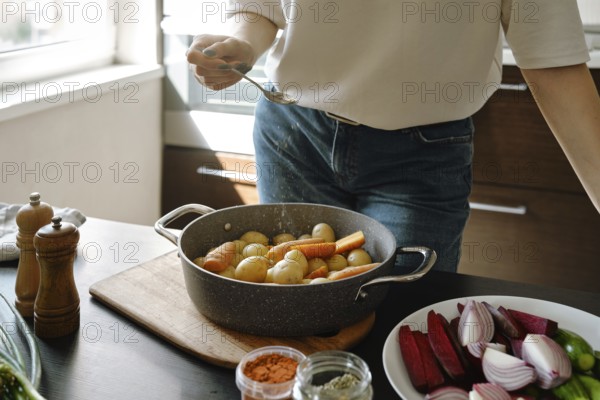 Unrecognizable woman sprinkles dried oregano on baby potatoes and carrots in a baking pan