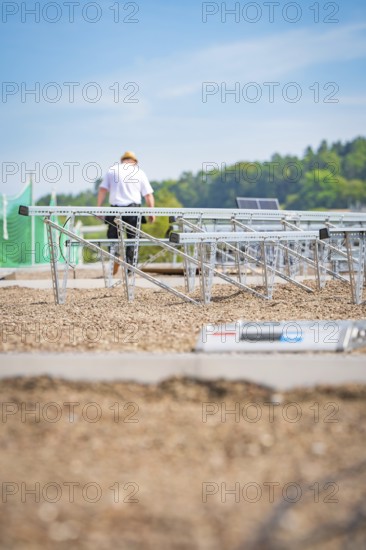 A worker assembles solar panel structures on a gravel roof under a blue sky, new park and ride