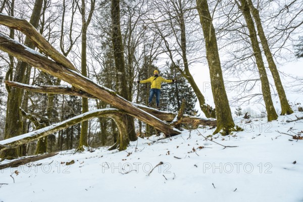 Man wearing a yellow jacket and hat balancing arms outstretched on a large fallen tree log covered
