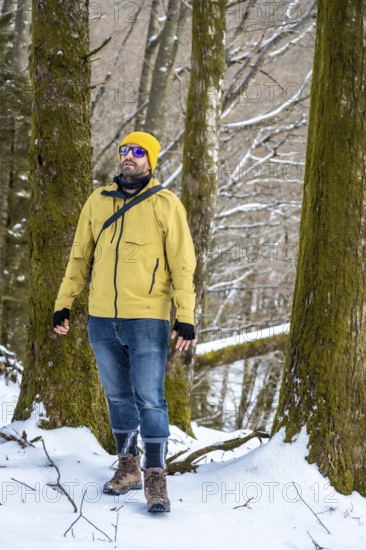Man wearing a yellow winter jacket, jeans, and hiking boots exploring a snowy forest landscape,