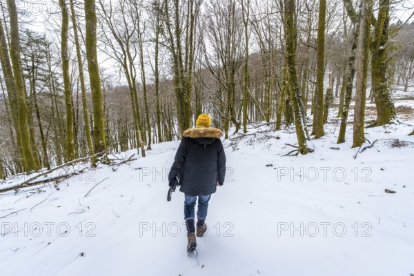 Person walking uphill in a winter forest, carrying a camera, wearing warm clothing with a yellow